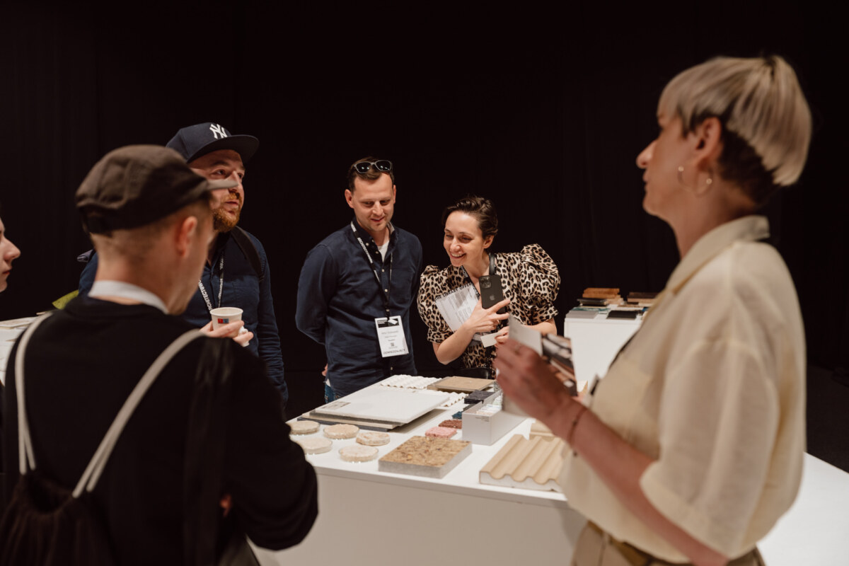 reportaz targi architektura architectatwork 2025 30 Eine Gruppe von fünf Personen steht um einen Tisch herum und präsentiert Proben und Materialien, während sie sich in einem Raum unterhalten. Eine Person gestikuliert, während die anderen aufmerksam zuhören.