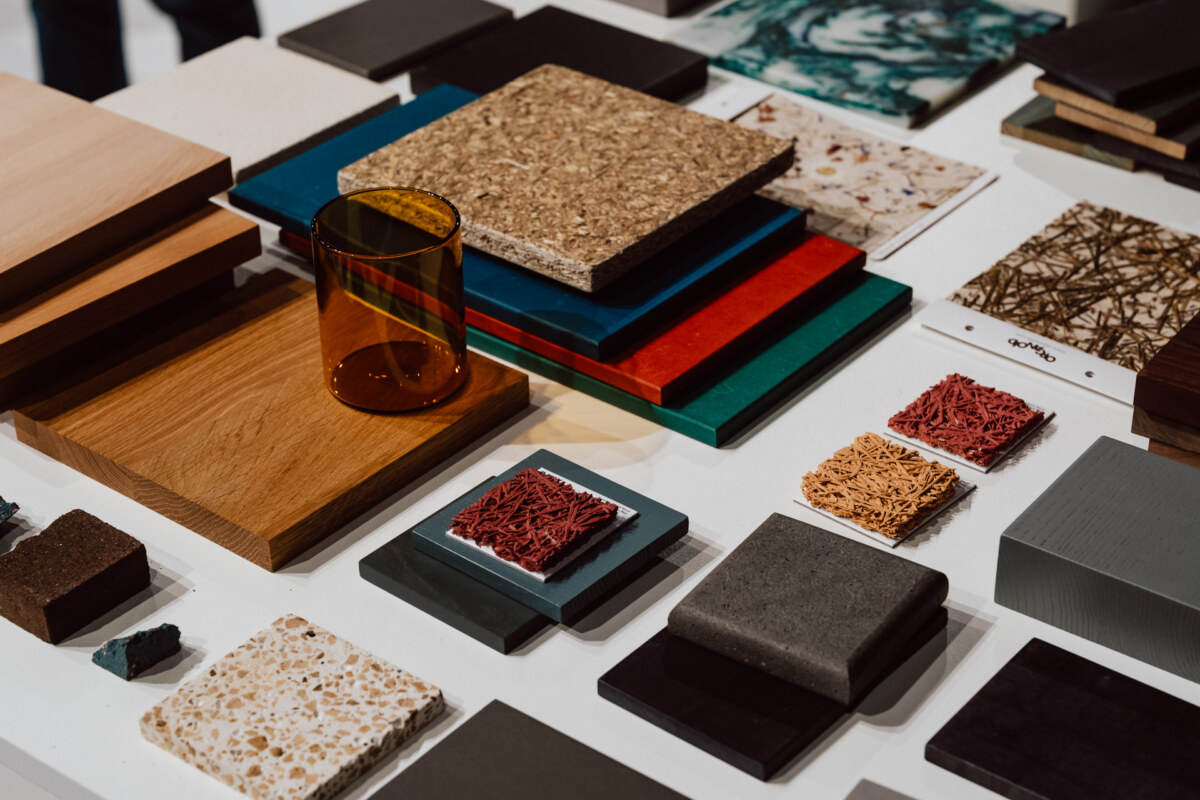 reportaz targi architektura architectatwork 2025 31 Eine Vielzahl von bunten Materialproben, darunter Holz, Stein, Kork und Kunststoff, sind ordentlich auf einem weißen Tisch neben einem durchsichtigen Braunglasbecher angeordnet.