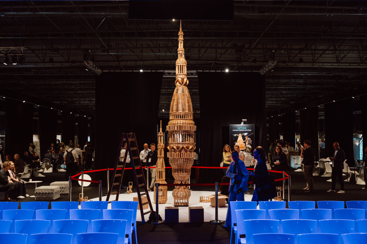 reportaz targi architektura architectatwork 2025 34 Eine hohe, verschlungene Holzskulptur steht in der Mitte der Ausstellungshalle, umgeben von einer roten Seilabsperrung und Menschen, die zusehen. Im Vordergrund stehen Reihen von leeren blauen Stühlen.