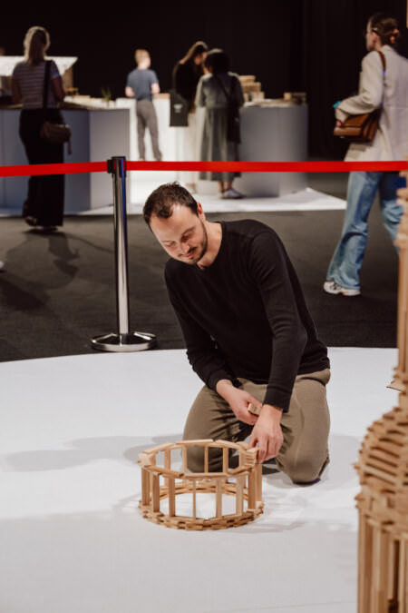 Ein Mann kniet auf dem Boden und lächelt, während er eine runde Holzstruktur aus Blöcken baut. Im Hintergrund, hinter einer roten Absperrung aus Seilen, sind weitere Personen zu sehen. 