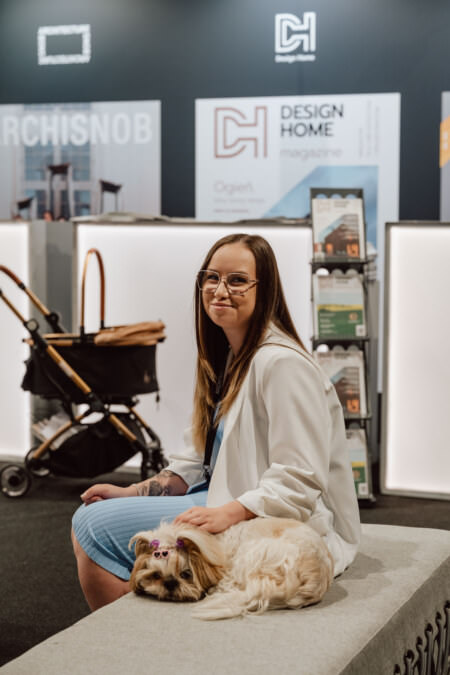 Eine Frau mit Brille und weißer Jacke sitzt auf einer Bank und lächelt, während ein kleiner Hund neben ihr liegt. Der Hund hat Schleifen in seinem Fell. Dahinter stehen Regale mit Zeitschriften und ein Einkaufswagen mit einem auffälligen 'Design Home'-Schriftzug.  