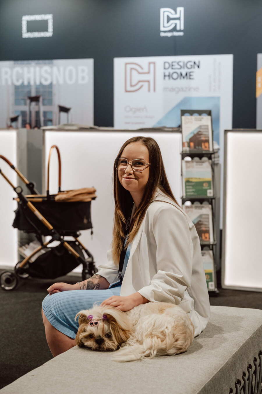 reportaz targi architektura architectatwork 2025 36 Eine Frau mit Brille und weißer Jacke sitzt auf einer Bank und lächelt, während ein kleiner Hund neben ihr liegt. Der Hund hat Schleifen in seinem Fell. Dahinter stehen Regale mit Zeitschriften und ein Einkaufswagen mit einem auffälligen 'Design Home'-Schriftzug.