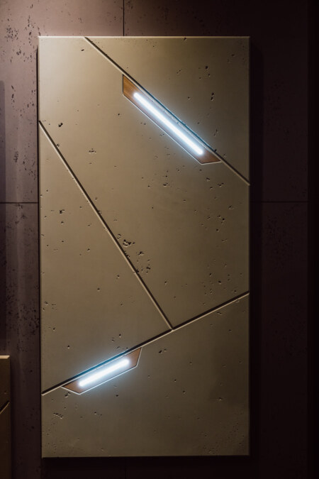 reportaz targi architektura architectatwork 2025 37 Ein rechteckiges beiges Paneel mit strukturierten diagonalen Linien und zwei eingebauten LED-Lichtstreifen, die ein kühles weißes Licht ausstrahlen, vor einer dunklen Wand.