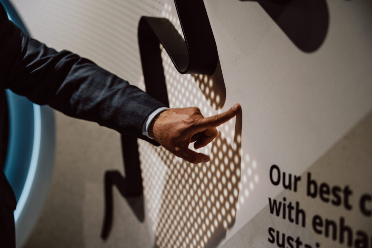 reportaz targi architektura architectatwork 2025 38 Die Person im Anzug zeigt auf den Text an der Wand, der teilweise sichtbar ist und lautet: "Unser bester K... mit enha... sustai...'. Das Licht erzeugt ein gepunktetes Schattenmuster an der Wand und auf der Hand.