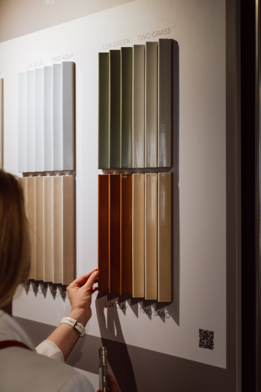Die Hand des Mannes greift nach einem rechteckigen Display mit Mustern von Keramikfliesen in verschiedenen Farben, darunter blau, grün, cremefarben, braun und beige, die an einer Wand mit Etiketten und einem kleinen QR-Code in der Ecke angebracht sind.