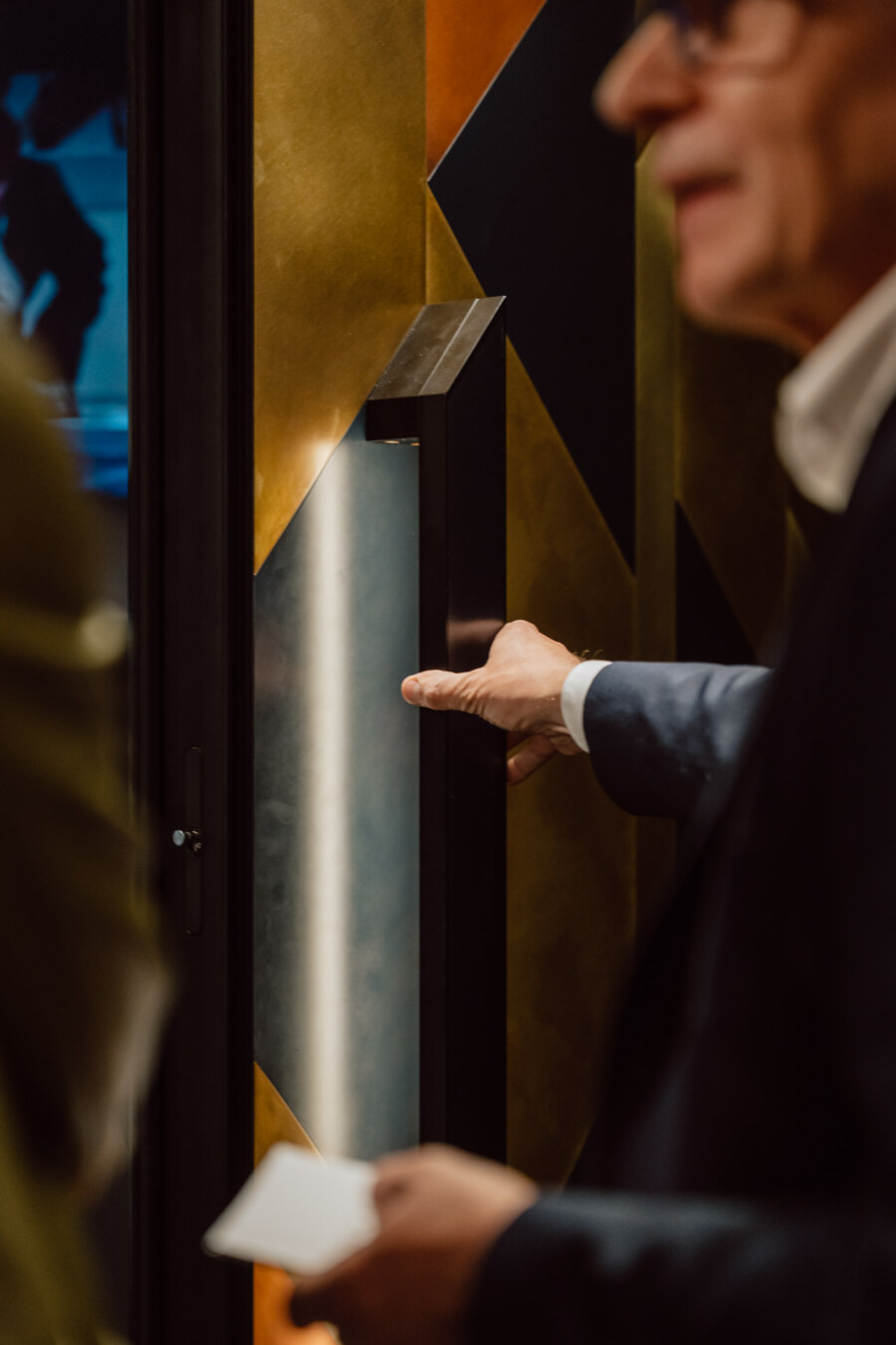 reportaz targi architektura architectatwork 2025 44 Eine Person im Anzug drückt einen Knopf oder Schalter auf einer Metalltafel, die an einer Wand mit geometrischen Mustern angebracht ist. Der Fokus liegt auf ihrer Hand, während ihr Gesicht unscharf ist.