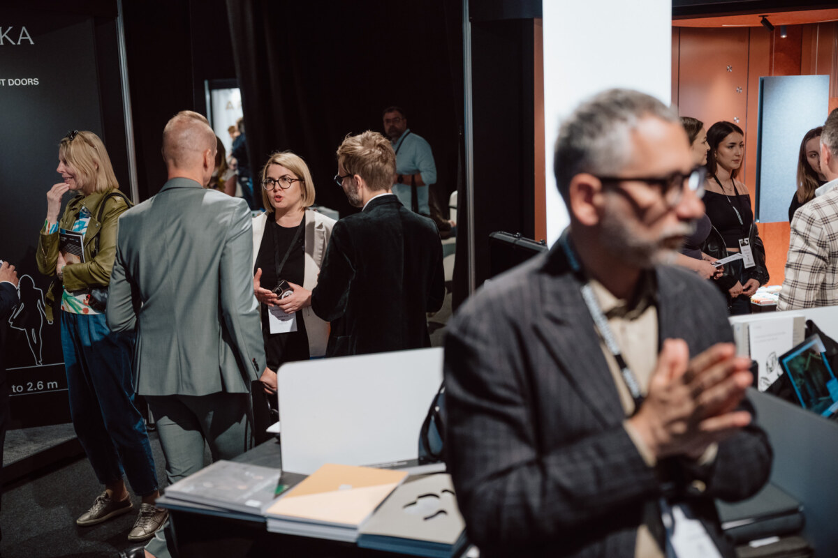 reportaz targi architektura architectatwork 2025 46 Eine Gruppe von Personen in Geschäftskleidung unterhält sich und knüpft Kontakte bei einer Veranstaltung oder Ausstellung in einem Raum, wobei Broschüren und Auslagen im Vordergrund zu sehen sind.