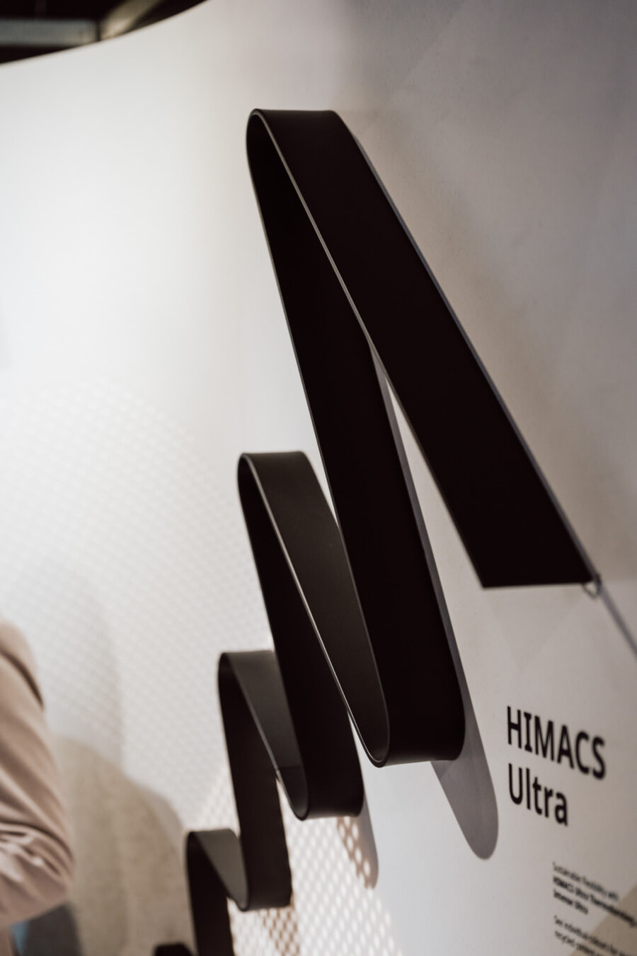 Die schwarze, bandartige Skulptur ist an einer weißen Wand neben der Aufschrift 'HIMACS Ultra' angebracht. Die Grafik bildet geschlungene, eckige Formen und am linken Rand ist ein Teil einer Person in heller Kleidung zu sehen. 