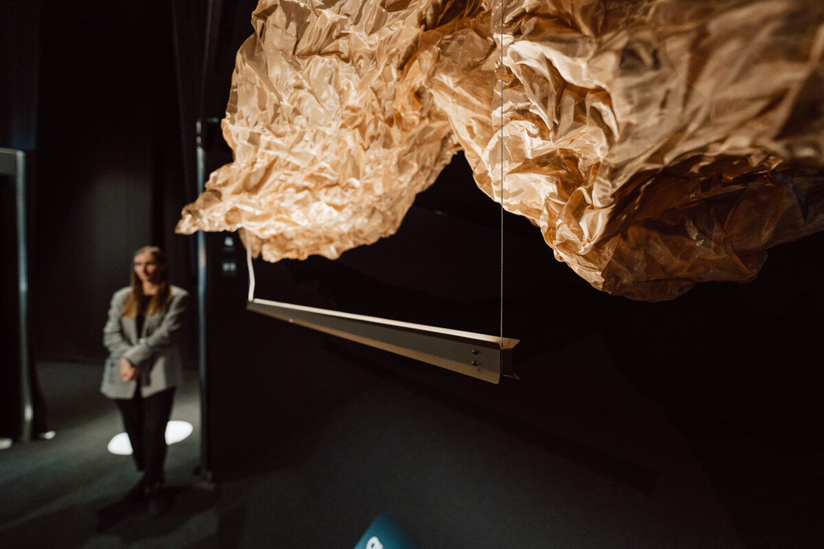 reportaz targi architektura architectatwork 2025 5 Die Frau steht in einem schwach beleuchteten Raum neben einem großen, zerknitterten braunen Tuch, das von der Decke herabhängt, mit einer kantigen Metallstange darunter.