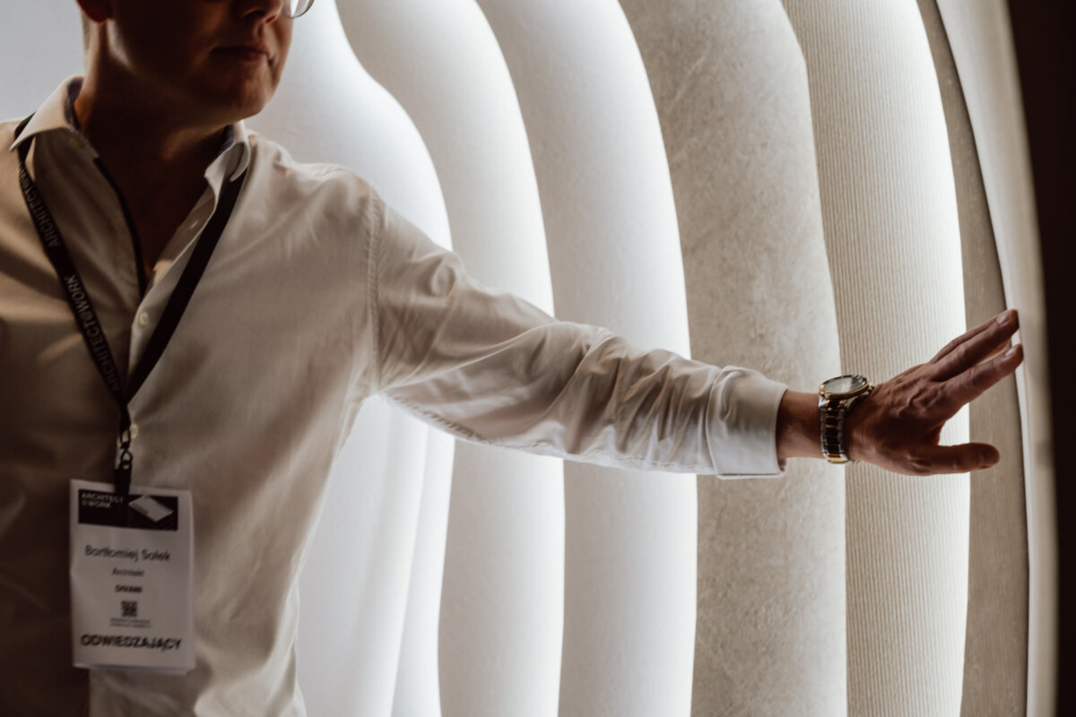 reportaz targi architektura architectatwork 2025 50 Eine Person mit weißem Hemd und Brille berührt mit ihrer rechten Hand eine gebogene, beleuchtete Wand. Ihr Ausweis ist teilweise sichtbar und das Licht erzeugt einen sanften, leuchtenden Effekt auf der Wand.