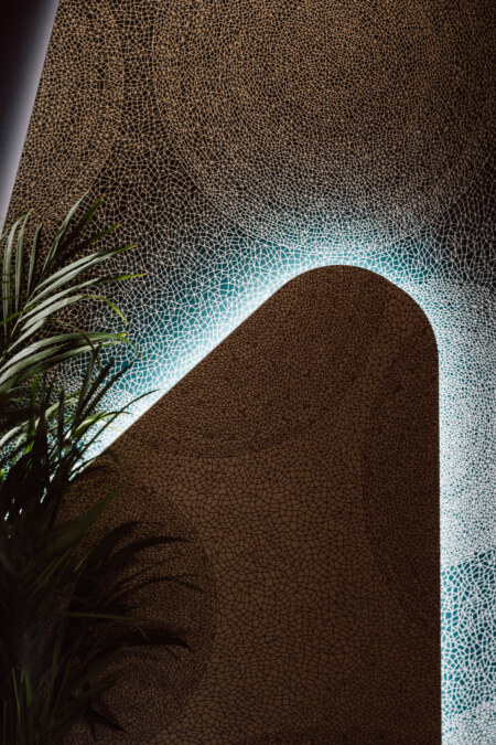 reportaz targi architektura architectatwork 2025 52 Eine strukturierte Mosaikwand mit einem Rundbogen wirft abstrakte Schatten. Blaues Licht hebt den Bogen hervor und eine grüne Pflanze ist in der unteren linken Ecke teilweise sichtbar.