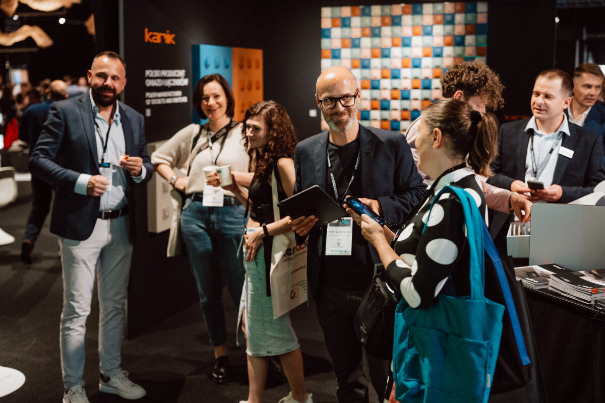 reportaz targi architektura architectatwork 2025 53 Eine Gruppe professionell gekleideter Personen steht während einer Geschäftsveranstaltung in einem Raum und unterhält sich, einige halten Telefone, Tablets oder Getränke in der Hand, während im Hintergrund Informationsdisplays und Broschüren zu sehen sind.