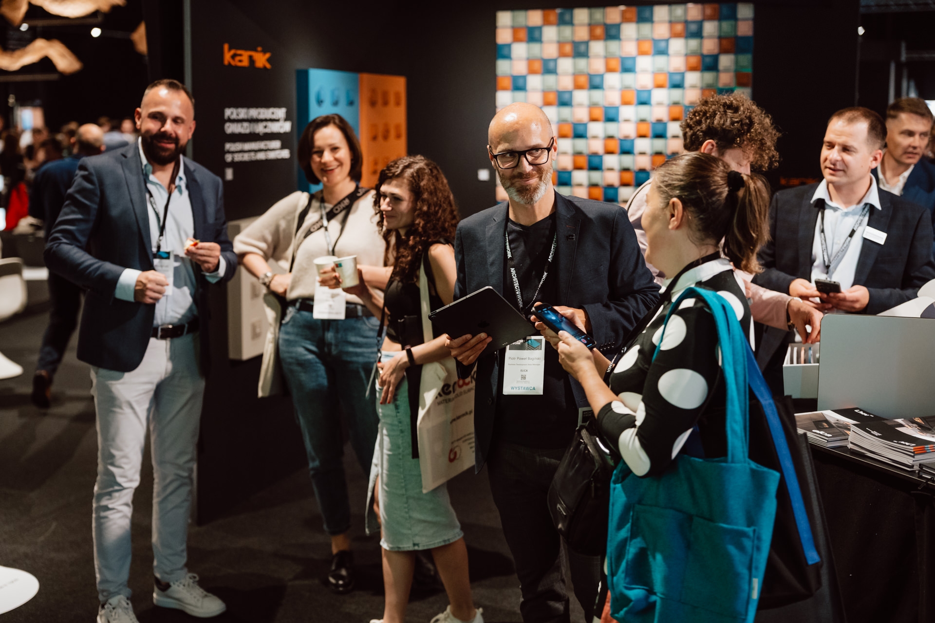 reportaz targi architektura architectatwork 2025 53 Trzy osoby siedzą razem na imprezie, uśmiechając się i trzymając drinki. Dwie noszą identyfikatory i pozują do kamery, wyglądając wesoło. W tle widać innych uczestników i ciemną przestrzeń wystawową.