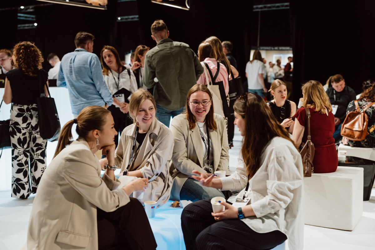 reportaz targi architektura architectatwork 2025 56 Eine Gruppe von Menschen sitzt und steht im Gespräch während einer lebhaften Veranstaltung in dem Raum. Vier Frauen im Vordergrund sitzen lächelnd und plaudernd auf weißen Bänken, während sich andere im Hintergrund unter hellen Lichtern mischen.