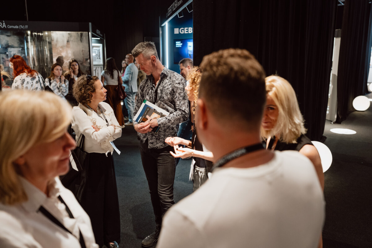 reportaz targi architektura architectatwork 2025 58 Eine Gruppe von Menschen steht während einer Veranstaltung oder Ausstellung in einem Raum und unterhält sich. Einige halten Broschüren in der Hand und im Hintergrund sind Messestände mit Schildern und Lichtern zu sehen. Die Umgebung ist lebhaft und professionell.