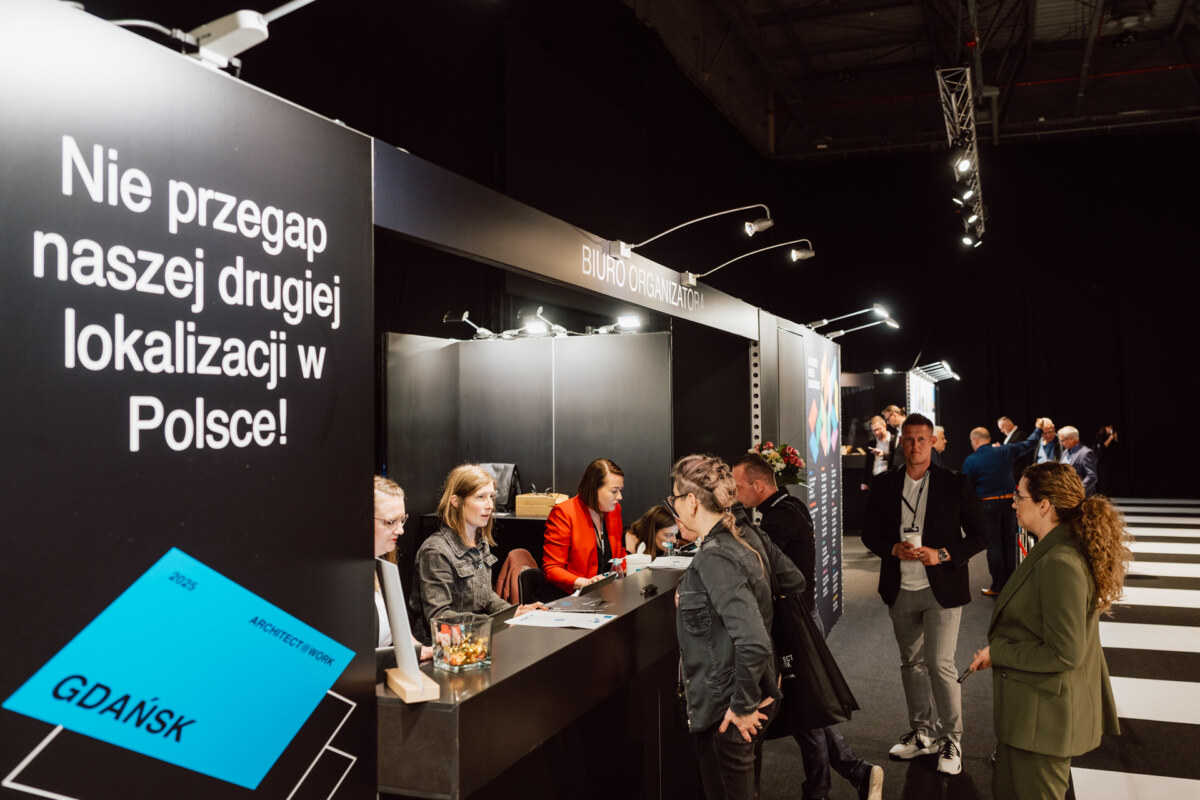 reportaz targi architektura architectatwork 2025 59 Menschen unterhalten sich an einem modernen Informationsstand in einem großen Veranstaltungsraum. Ein deutliches schwarzes Schild in polnischer Sprache ist zu sehen und mehrere Mitarbeiter sitzen an der Theke, während die Teilnehmer in der Nähe stehen und sich unterhalten.