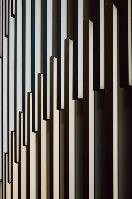 reportaz targi architektura architectatwork 2025 6 Vertikale, rechteckige Leuchten in unterschiedlichen Höhen sind an der Wand angebracht. So entsteht ein modernes geometrisches Muster mit abwechselnden Schattierungen von Weiß und Dunkelbraun.