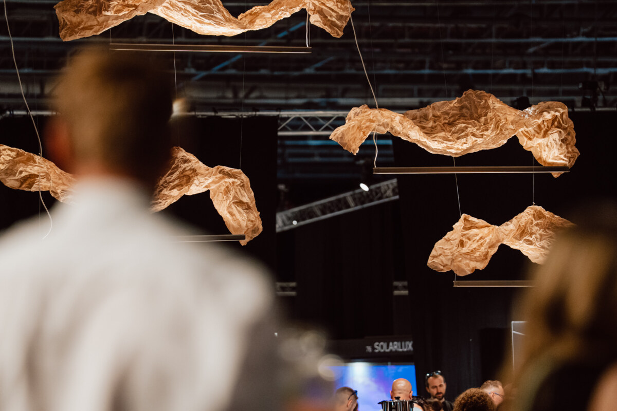 reportaz targi architektura architectatwork 2025 60 Hängende abstrakte Skulpturen aus zerknittertem, strukturiertem Material werden bei einer Indoor-Kunstausstellung über die Menge projiziert. Der Vordergrund ist leicht verschwommen und zeigt Menschen, die die Installation beobachten.