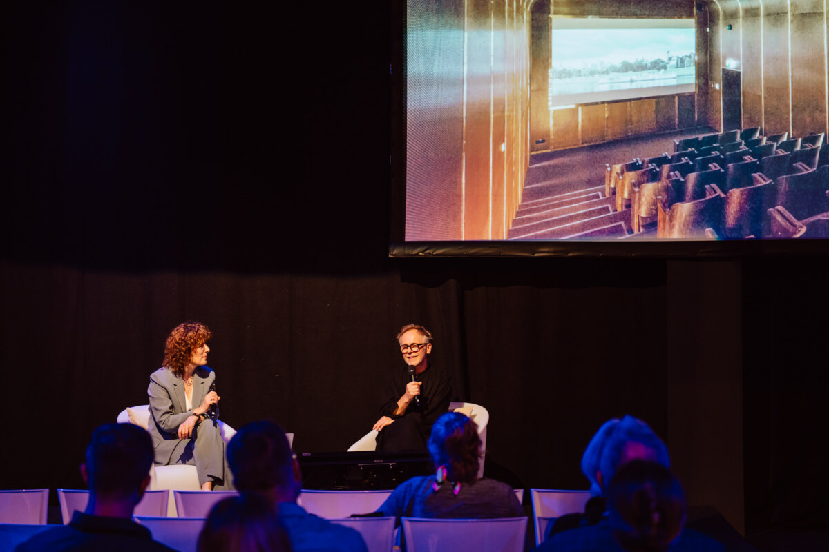 reportaz targi architektura architectatwork 2025 64 Zwei Personen sitzen auf der Bühne und diskutieren mit Mikrofonen vor einem Publikum. Ein großer Bildschirm über ihnen zeigt das Bild eines leeren Theaters mit Sitzreihen vor einem großen Fenster.