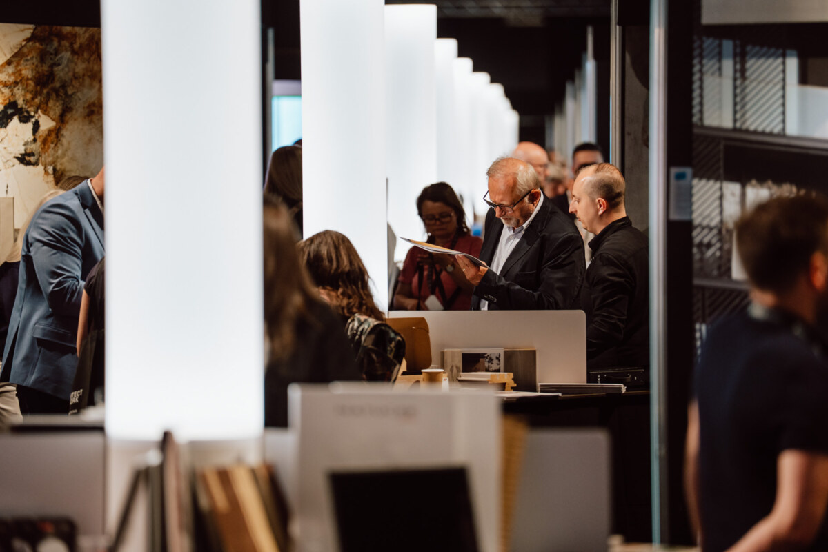 reportaz targi architektura architectatwork 2025 67 Eine Gruppe von Menschen, einige stehend und einige lesend, versammelt sich in einem modernen, gut beleuchteten Innenraum mit hohen weißen Leuchttafeln und verschiedenen Displays. Die Atmosphäre erinnert an eine professionelle Veranstaltung oder eine Ausstellung.