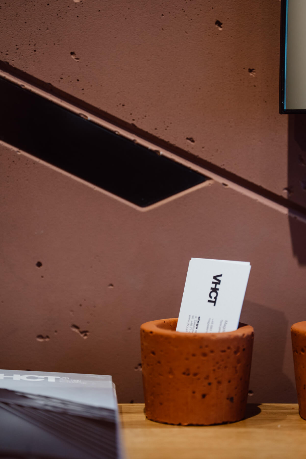 Eine weiße Visitenkarte mit dem Wort 'VHCT' steht senkrecht in einem kleinen, strukturierten, orange-braunen Topf auf einer hölzernen Oberfläche vor einer violetten Wand im Industriestil mit diagonalen schwarzen Akzenten.