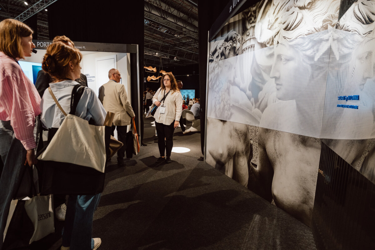 reportaz targi architektura architectatwork 2025 7 Eine Gruppe von Menschen steht in dem Ausstellungsraum und hört einer Frau in einer weißen Jacke zu, die neben einer Ausstellung klassischer Statuen spricht. Der Raum ist schwach beleuchtet, modern dekoriert und mit Informationstafeln versehen.