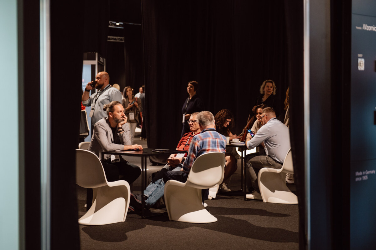 reportaz targi architektura architectatwork 2025 74 Eine Gruppe von Menschen sitzt an runden Tischen und unterhält sich in einem schwach beleuchteten Raum mit schwarzen Vorhängen. Einige sind in ein Gespräch vertieft, während andere zuhören. Moderne weiße Stühle umgeben die Tische. Im Hintergrund sind weitere Personen zu sehen.