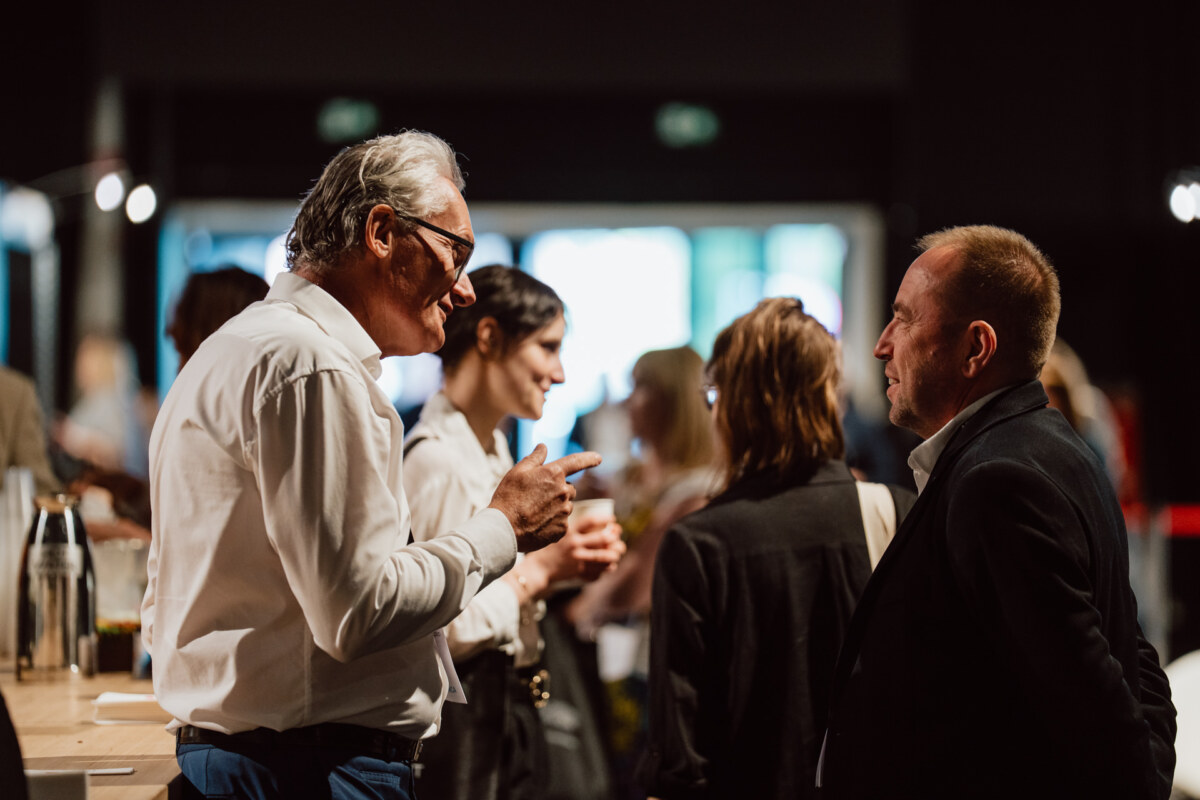 reportaz targi architektura architectatwork 2025 77 Zwei Männer unterhalten sich bei einem gesellschaftlichen Ereignis, umgeben von anderen, die in einem gut beleuchteten Innenraum diskutieren. Die Atmosphäre wirkt lebendig und professionell.