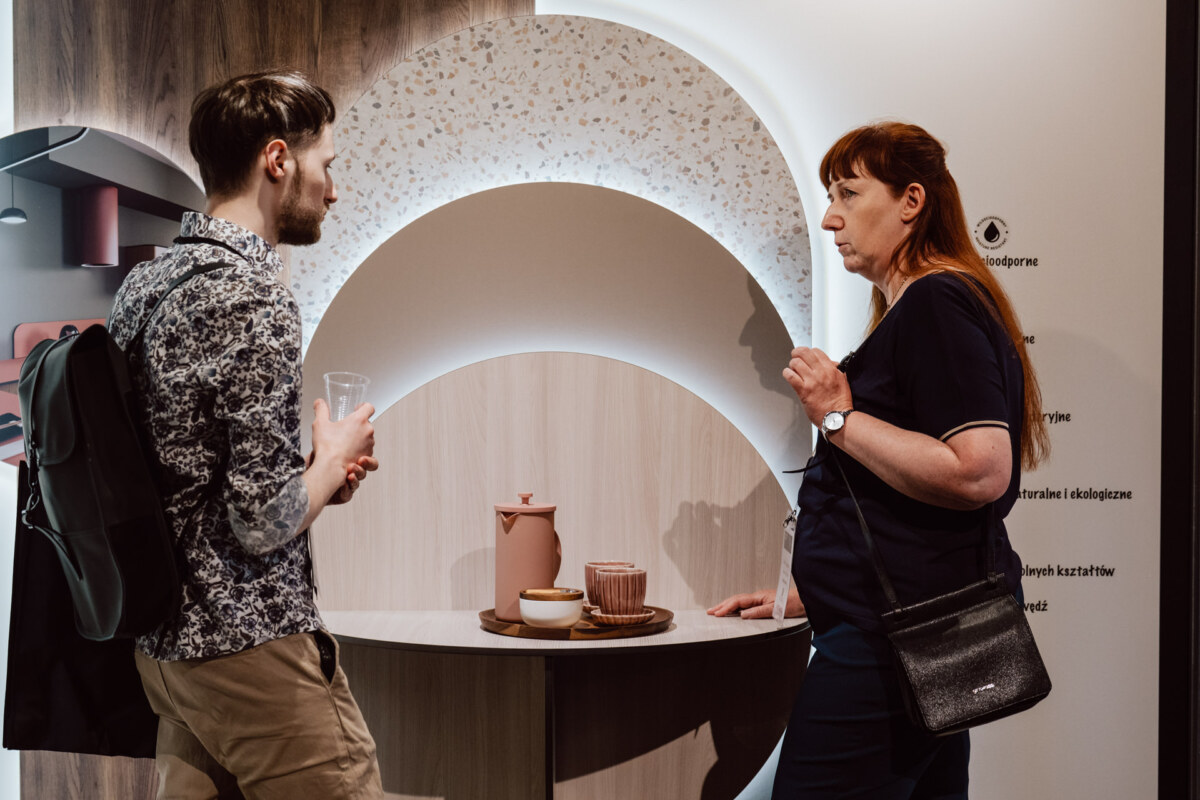 reportaz targi architektura architectatwork 2025 78 Ein Mann mit einem Rucksack und eine Frau mit einer Umhängetasche stehen sich gegenüber und unterhalten sich in der Nähe eines runden Regals mit Keramikgeschirr und einem Krug in einem modernen Interieur.