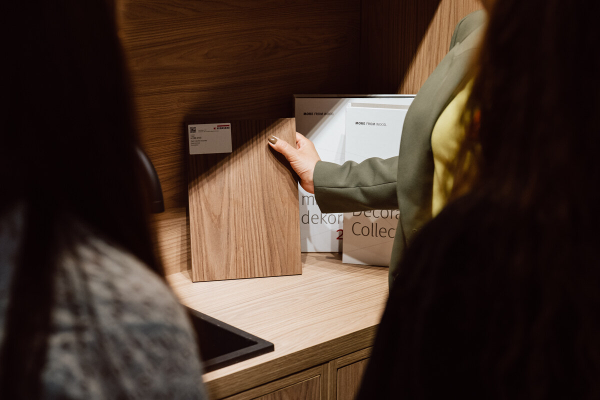 reportaz targi architektura architectatwork 2025 79 Eine Person hält ein Holzbrett mit Mustern in der Nähe der Küchentheke, während zwei andere Personen sie beobachten. Dekorative Musterbücher sind an einer Holzwand im Hintergrund aufgehängt.