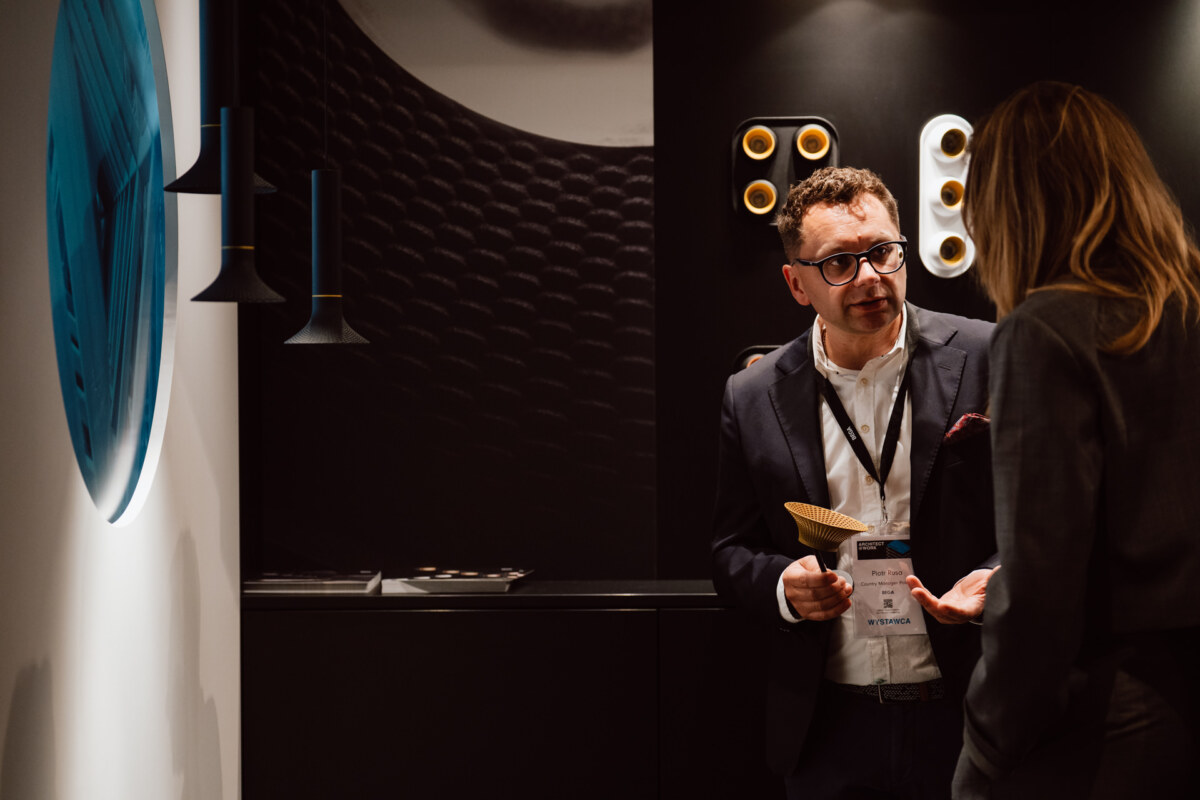reportaz targi architektura architectatwork 2025 80 Ein Mann im Anzug und mit Brille spricht mit einer Frau in einem schwach beleuchteten, modernen Interieur mit abstrakten Wandgrafiken und dekorativen Lichtern im Hintergrund.
