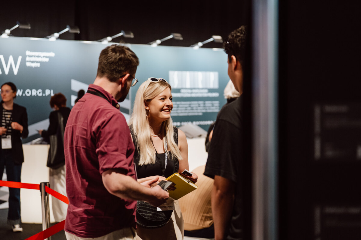 reportaz targi architektura architectatwork 2025 82 Eine lächelnde Frau mit blondem Haar unterhält sich bei einer Indoor-Veranstaltung mit zwei Männern, die Dokumente halten. Im Hintergrund ist ein Stand mit Text und Grafiken zu sehen. Rote Seile trennen den Bereich ab.