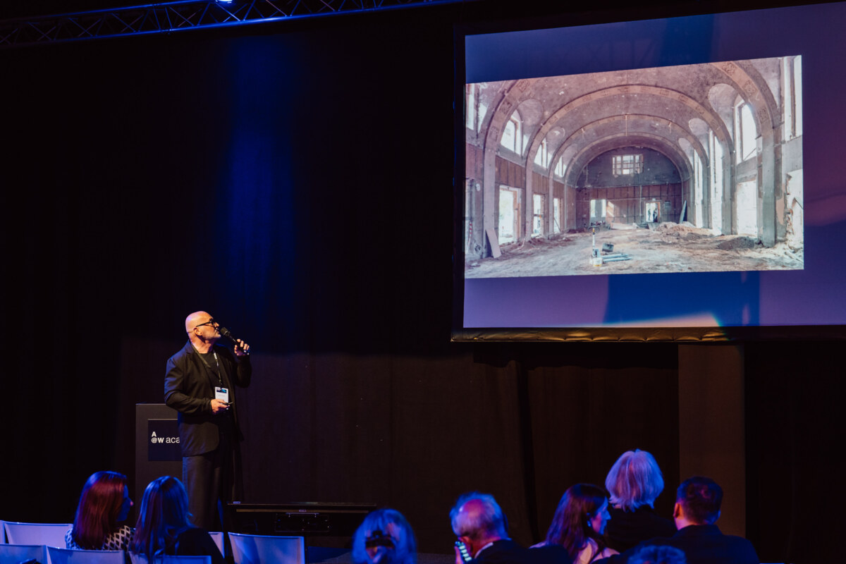 reportaz targi architektura architectatwork 2025 83 Ein Mann im Anzug trinkt aus einer Flasche, steht auf der Bühne und wendet sich an das Publikum. Ein großer Bildschirm hinter ihm zeigt ein Foto des leeren, gewölbten und teilweise renovierten Innenraums des Gebäudes.
