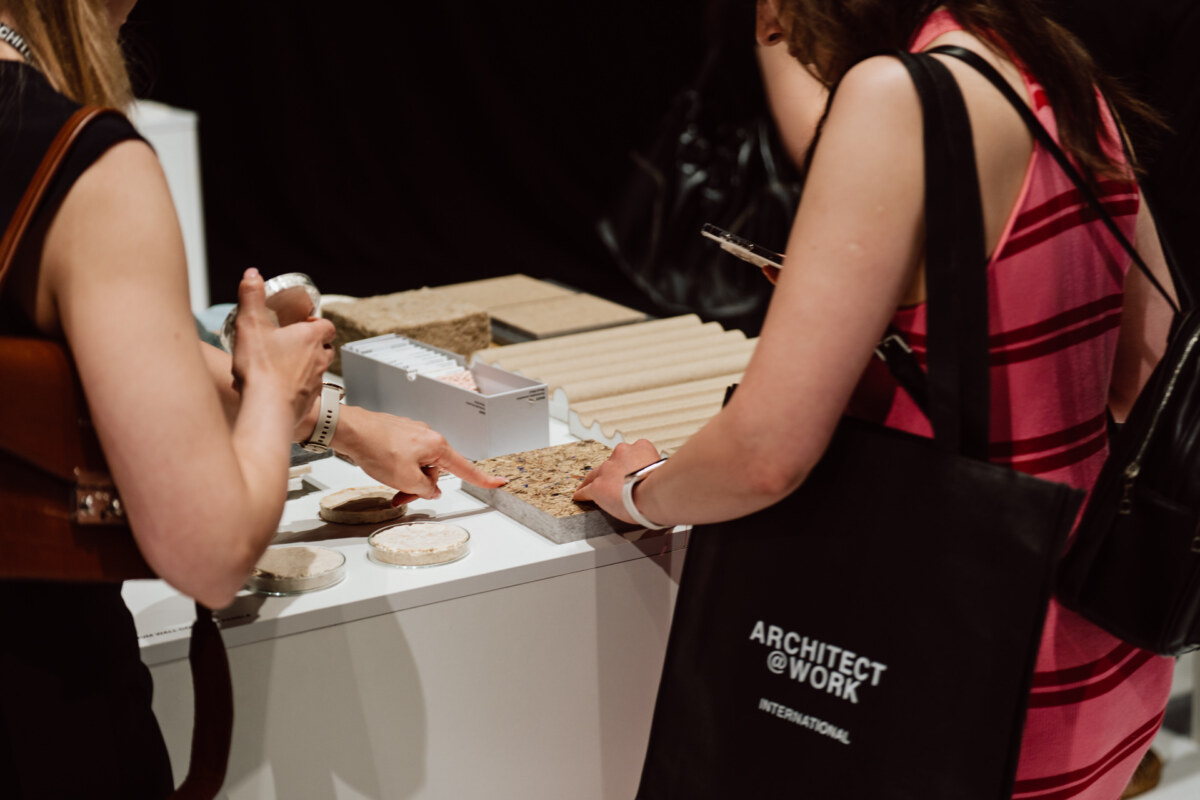 reportaz targi architektura architectatwork 2025 86 Zwei Frauen betrachten und berühren Proben von Baumaterialien auf einem Ausstellungstisch. Eine von ihnen zeigt auf ein strukturiertes Quadrat, während die andere eine schwarze Tasche mit der Aufschrift 'ARCHITECT @ WORK INTERNATIONAL' hält.