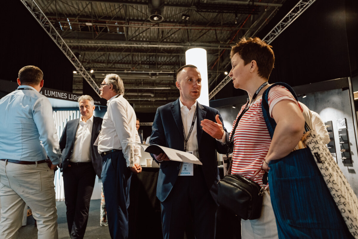 reportaz targi architektura architectatwork 2025 9 Eine Gruppe von Personen in Geschäftskleidung unterhält sich auf einer Messe oder Konferenz. Ein Mann hält Dokumente in der Hand, während er mit einer Frau in einem gestreiften Hemd und einer blauen Tasche spricht. Im Hintergrund sind Auslagen und Oberlichter zu sehen.