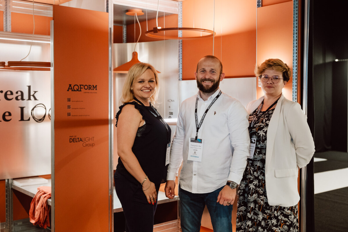 reportaz targi architektura architectatwork 2025 91 Drei Personen stehen lächelnd vor einem orangefarbenen Messestand mit moderner Beleuchtung und Beschilderung. Sie tragen Geschäftskleidung und Abzeichen, was darauf hindeutet, dass sie sich auf einer professionellen Veranstaltung oder Konferenz befinden.