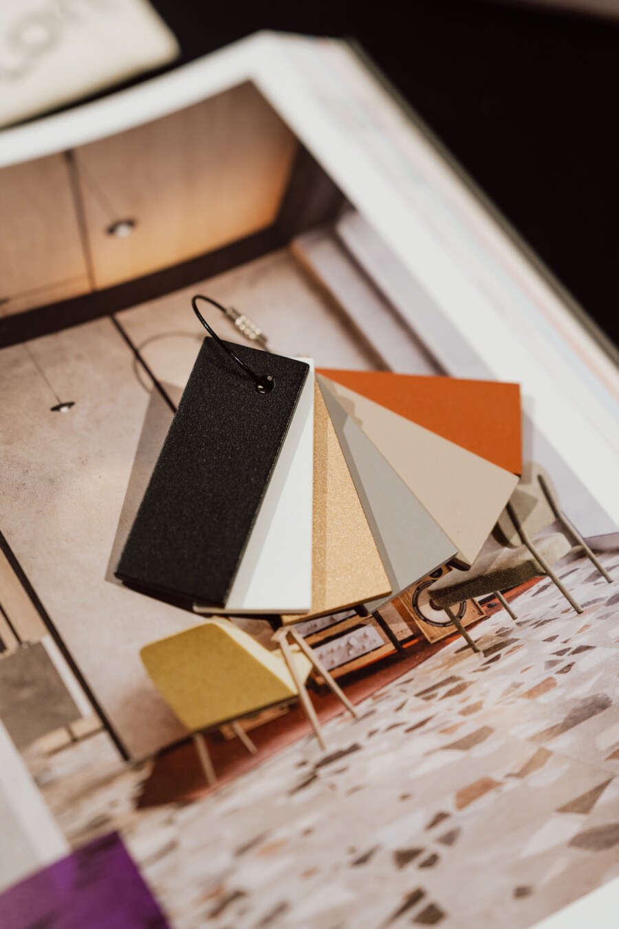 Eine Reihe von Farbmustern in Schwarz, Grau, Beige, Weiß und Orange liegt auf einem aufgeschlagenen Einrichtungsmagazin, das ein modernes Zimmer mit stilvollen Möbeln und gemustertem Bodenbelag zeigt.