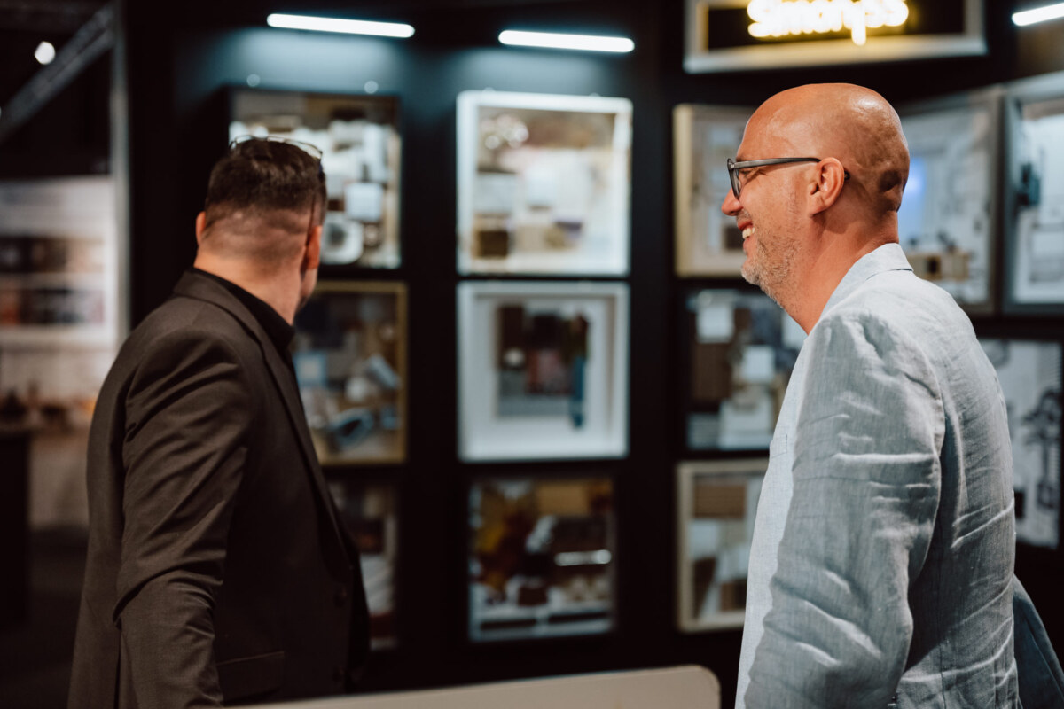 reportaz targi architektura architectatwork 2025 93 Zwei Männer in Anzügen stehen in einem Raum und betrachten gerahmte Kunstwerke oder Fotografien an der Wand. Einer von ihnen wendet sich der Ausstellung zu, während der andere, der eine Brille und eine hellblaue Jacke trägt, lächelt und zusieht. Es sieht aus wie eine Ausstellung.