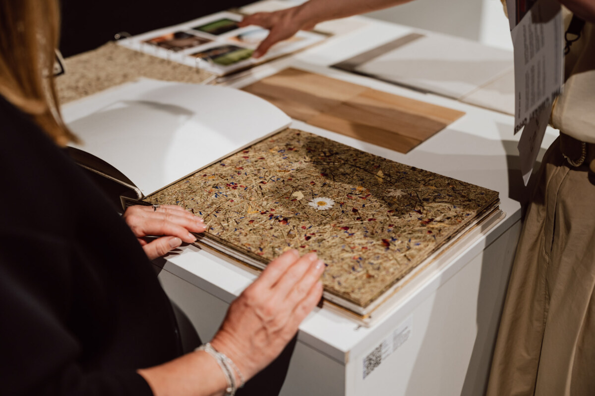 reportaz targi architektura architectatwork 2025 99 Zwei Personen betrachten ein Musterbuch mit einem strukturierten Material, in das getrocknete Blumen und Blätter eingebettet sind, während auf einem Tisch in der Nähe weitere Materialproben und Fotos zu sehen sind.