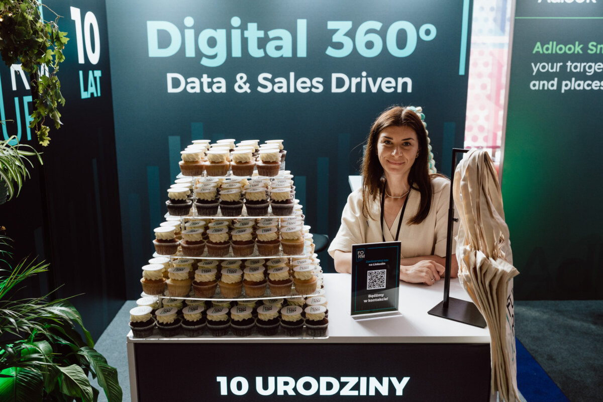 Kobieta stoi za ladą z piramidą małych słoiczków, prawdopodobnie z deserami, na stoisku z napisem "Digital 360° Data & Sales Driven". Na ladzie widoczny jest kod QR i napis "10 URODZINY".