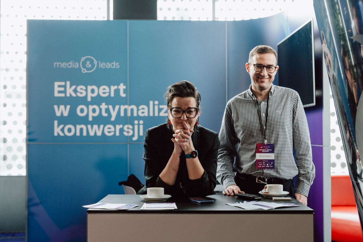 Dwie osoby stoją przy stoisku konferencyjnym z niebieskim napisem "Ekspert w optymalizacji konwersji". Uśmiechają się, a na stoliku przed nimi leżą kubki, papiery i identyfikatory.