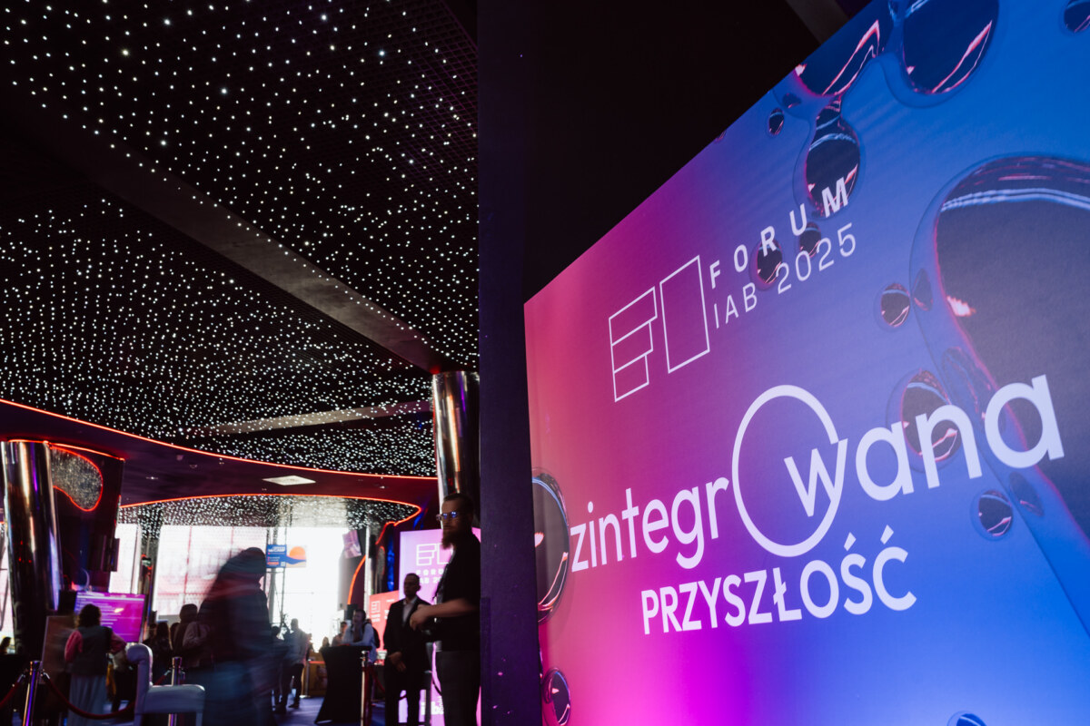 Ludzie gromadzą się w nowoczesnym, słabo oświetlonym pomieszczeniu z sufitem przypominającym gwiazdę na Forum IAB 2025. Kolorowy napis w języku polskim głosi "Zintegrowana Przyszłość" w pobliżu wejścia na wydarzenie.