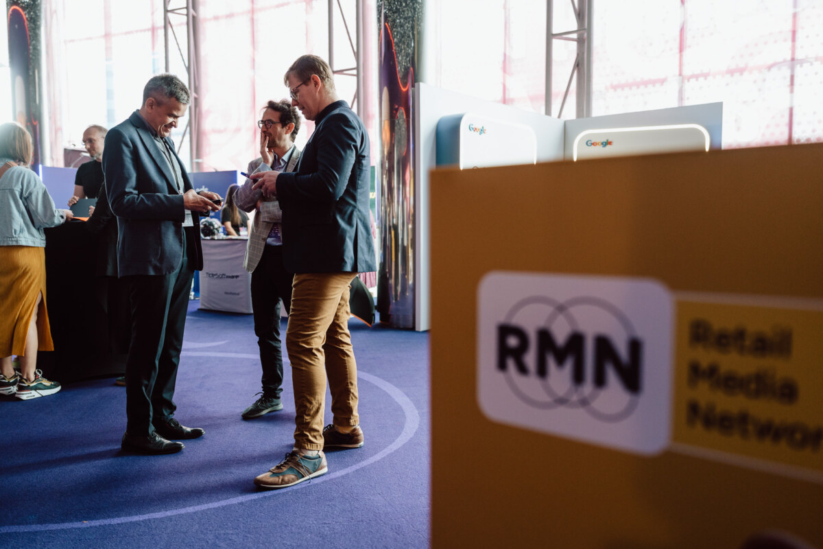 Ludzie nawiązujący kontakty podczas wydarzenia z żółtym znakiem "RMN Retail Media Network" na pierwszym planie i stoiskami Google widocznymi w tle.
