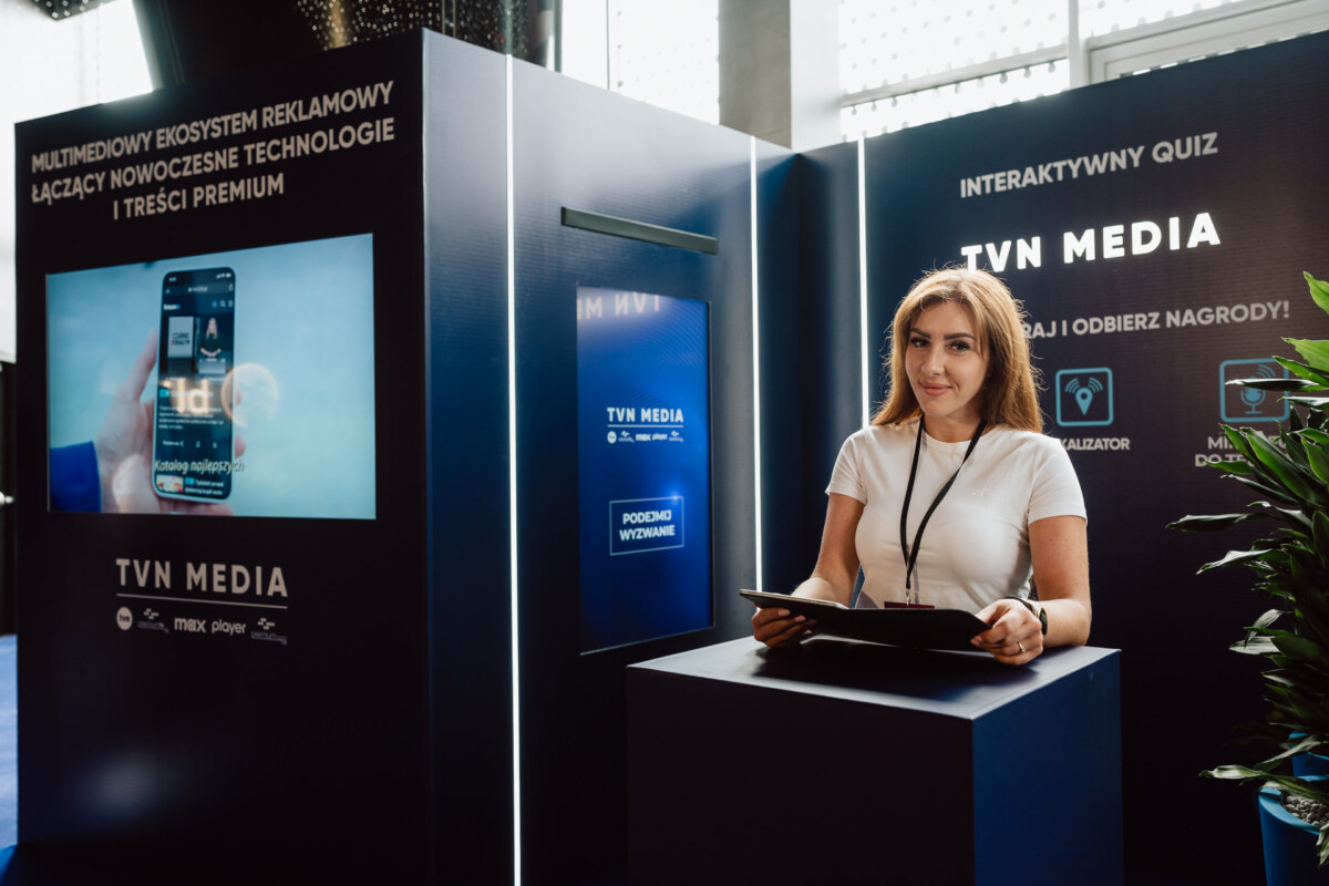 Kobieta stoi przy eleganckiej, nowoczesnej ladzie trzymając tablet przed stoiskami promocyjnymi TVN Media, na których znajdują się interaktywne znaki quizowe i wyświetlacze technologii reklamowych na imprezie wewnętrznej.