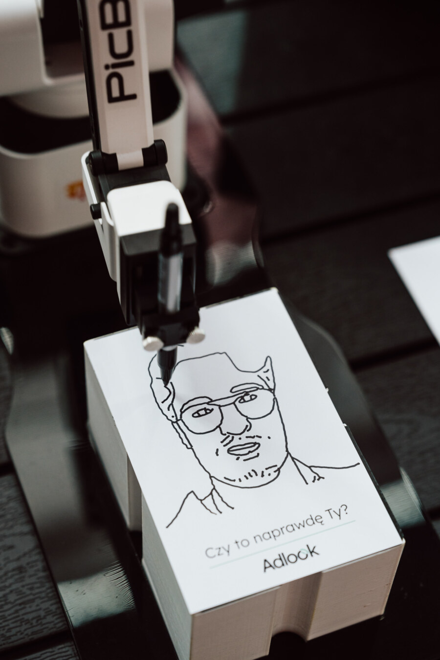Robotyczne ramię rysuje na kartce papieru portret mężczyzny w okularach i z brodą. Tekst na kartce brzmi "Czy to naprawdę Ty? Adlook.