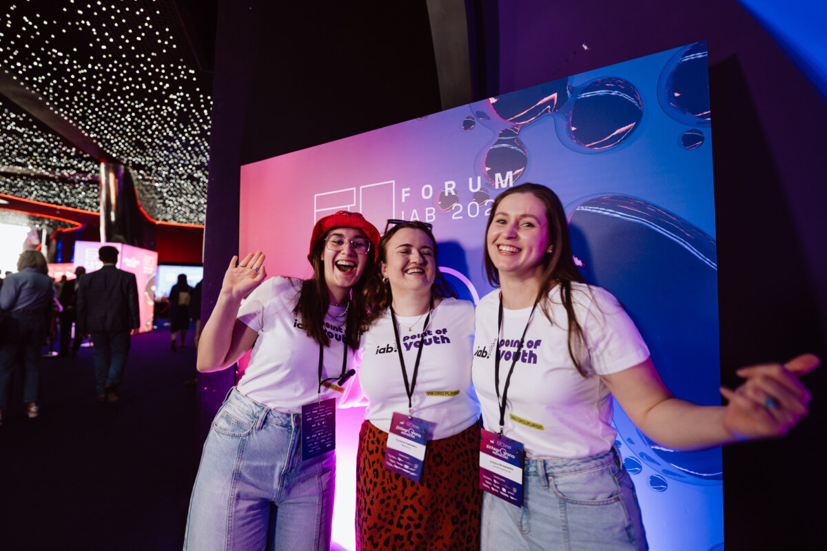 Trzy uśmiechnięte młode kobiety ubrane w koszulki "lab of youth" i plakietki wydarzenia pozują razem przed kolorowym napisem "Forum Lab 2023" w pomieszczeniu z wiszącymi nad głową światłami.