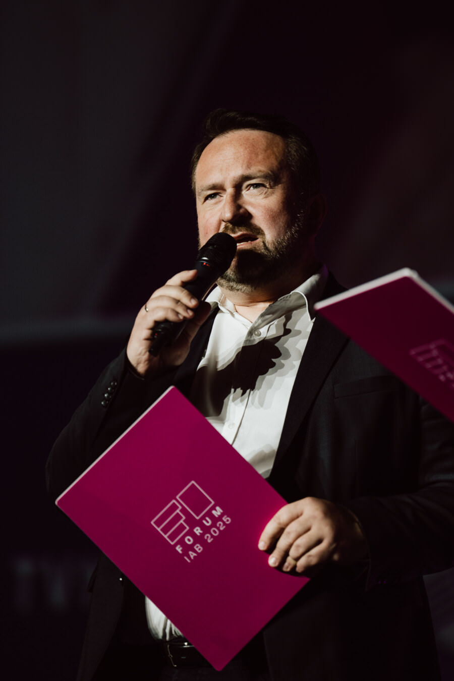 Ein Mann im Anzug hält ein Mikrofon und eine rosafarbene Aktentasche mit der Aufschrift 'IAB Forum 2023' und spricht auf der Bühne bei dramatischer Beleuchtung.