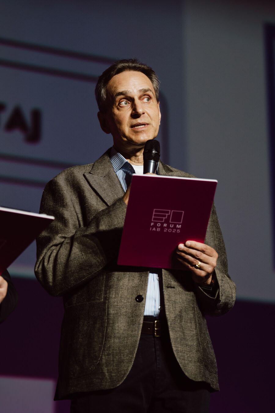 Ein Mann in einer grauen Jacke hält ein Mikrofon und eine purpurrote Aktentasche mit der Aufschrift 'IAB Forum 2023' in der Hand und spricht auf der Bühne, während das Logo im Hintergrund projiziert wird.