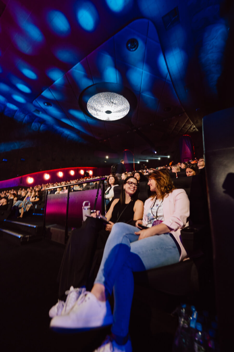 Zwei Personen sitzen und unterhalten sich in einem dunklen, modernen Theater, in dem viele andere um sie herum sitzen. Blaues und violettes Licht schafft eine lebendige Atmosphäre und ein großer Kronleuchter hängt von der Decke über dem Publikum. 