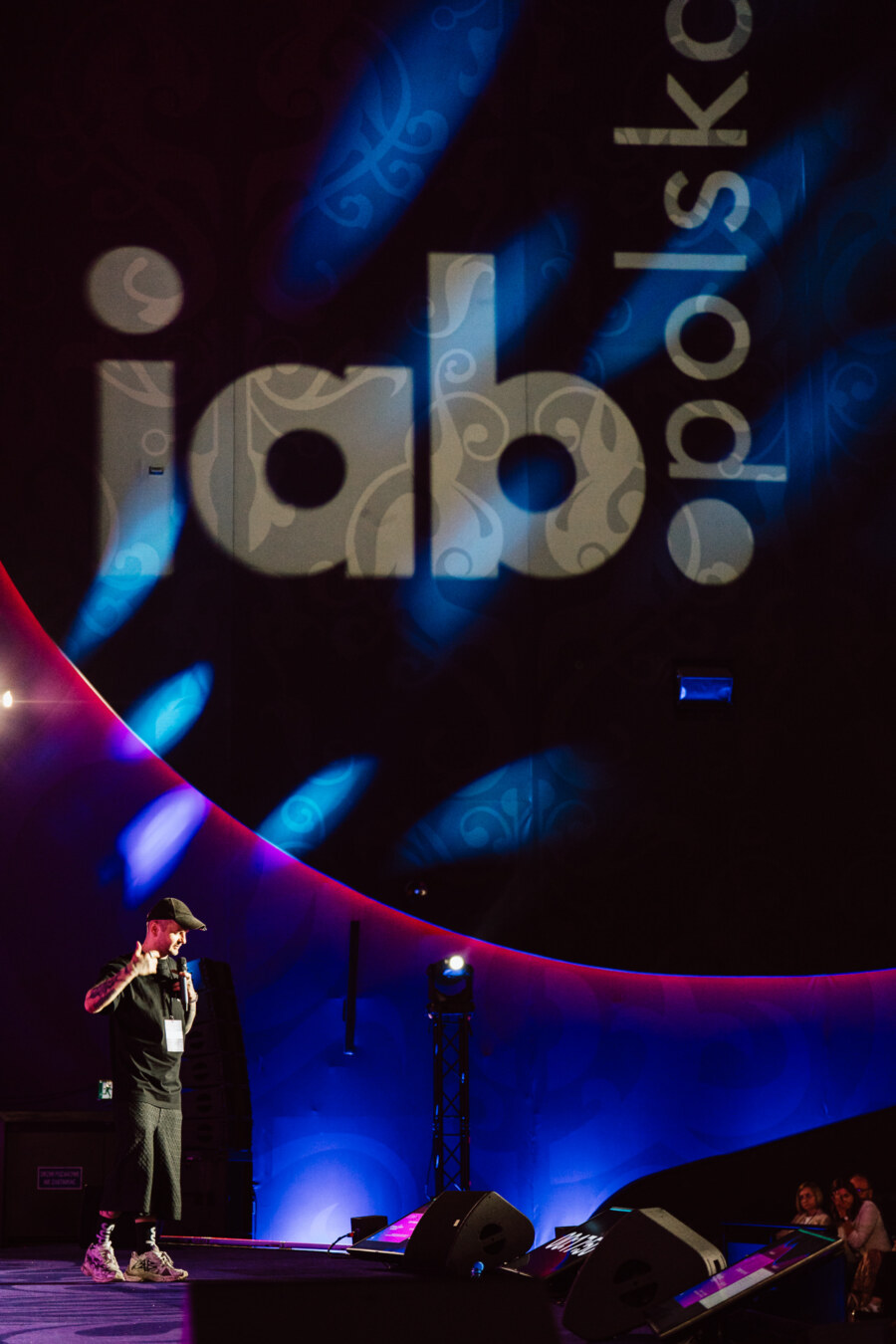 Mężczyzna w czapce i swobodnym ubraniu przemawia na scenie z dramatycznym oświetleniem. Duże, podświetlone logo "iab polska" jest wyświetlane na zakrzywionej ścianie za nim. Członkowie publiczności są częściowo widoczni w prawym dolnym rogu.